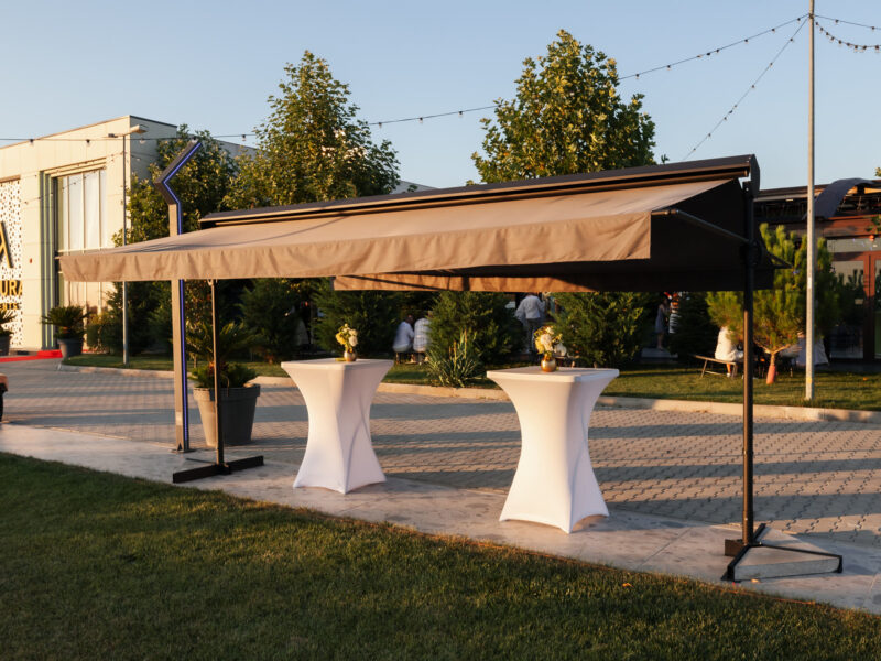 Marchiză bej de 5 pe 3 metri, deschisă peste două mese înalte de cocktail, la apus