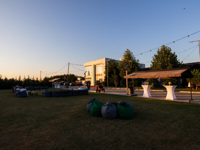 Zonă de eveniment în aer liber la apus, cu marchize deschise, mese înalte și zonă de relaxare pe gazon